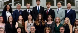 Eine Gruppe von Menschen, die feierlich gekleidet sind, sitzen und stehen in Reihen, wahrscheinlich für ein Foto. Einige lächeln. Sie befinden sich in einem Innenbereich mit einer weißen Wand und Glasfenstern.