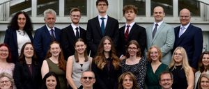 Eine Gruppe von Menschen, die feierlich gekleidet sind, sitzen und stehen in Reihen, wahrscheinlich für ein Foto. Einige lächeln. Sie befinden sich in einem Innenbereich mit einer weißen Wand und Glasfenstern.