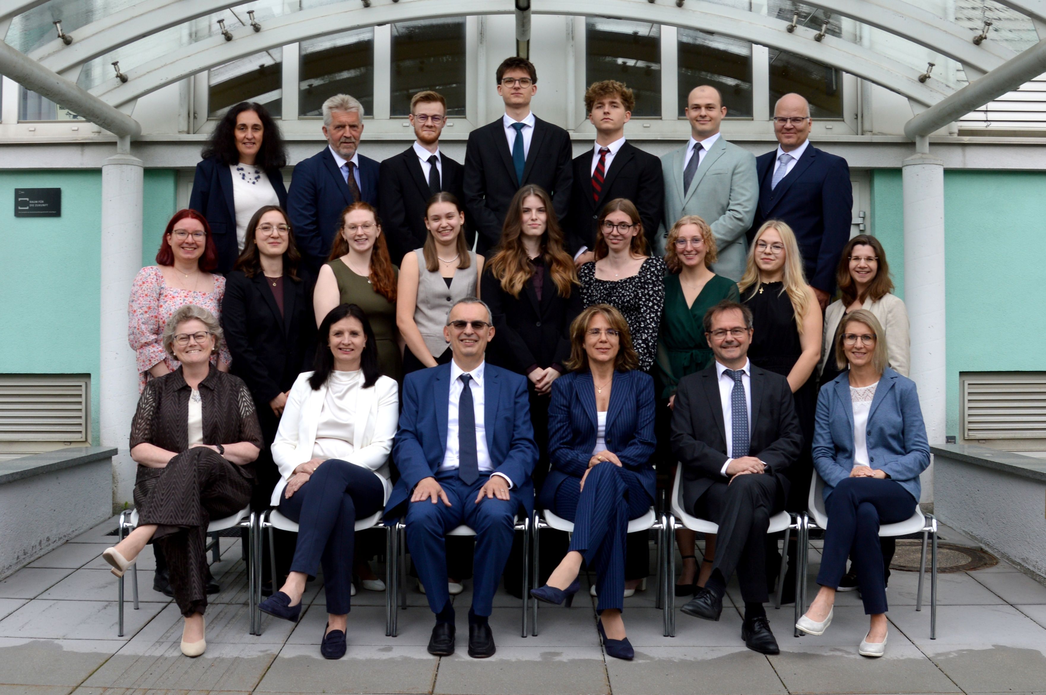 Eine Gruppenaufnahme einer vielfältigen Gruppe von Fachleuten und Studenten in formeller Kleidung, die vor einem modernen Gebäude sitzen und stehen.