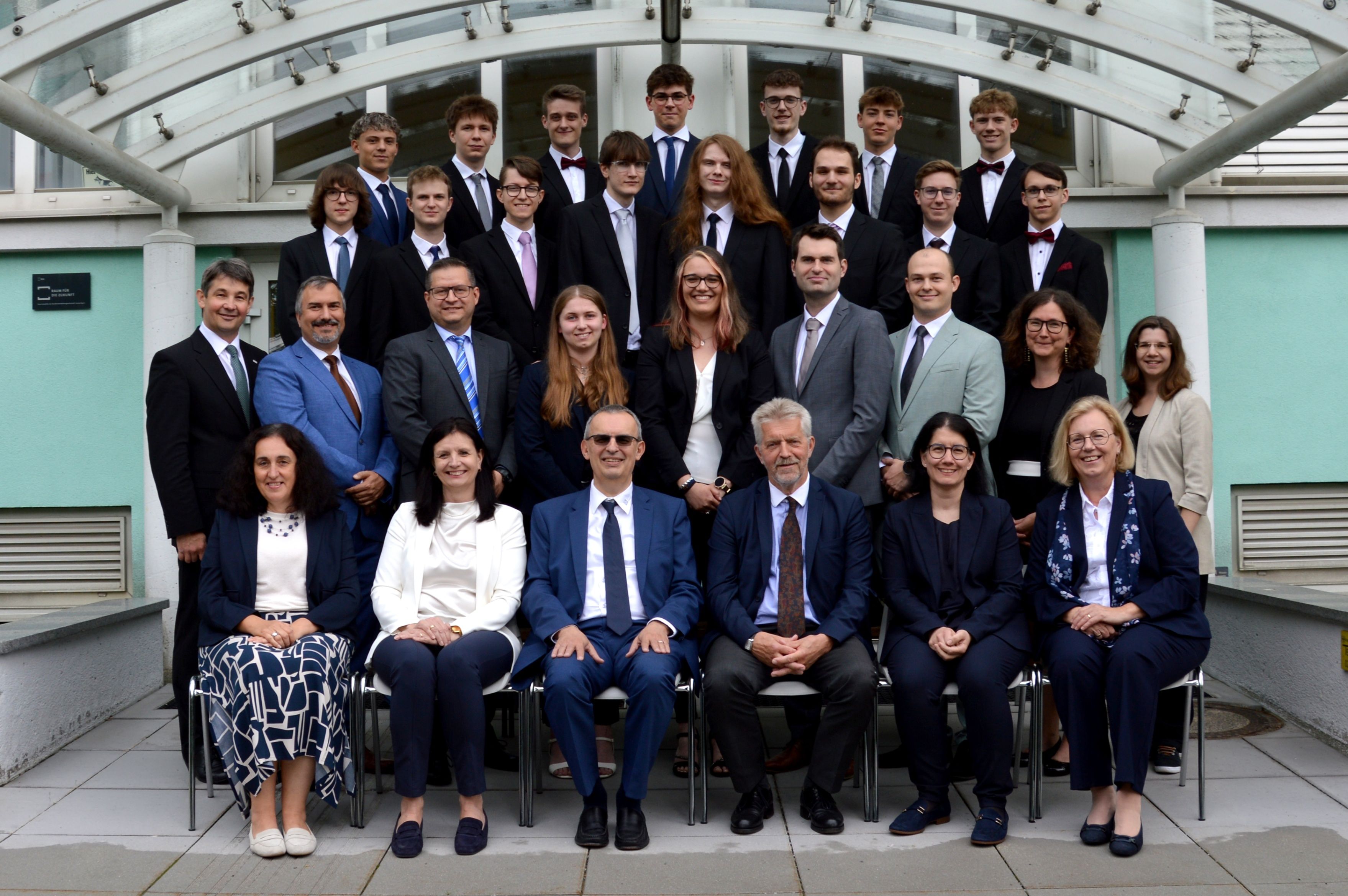 Ein Gruppenfoto zeigt Menschen in formeller Kleidung, darunter Männer in Anzügen und Krawatten und Frauen in Business-Anzügen, die zusammenstehen und sitzen. Sie alle lächeln und scheinen bei einer Veranstaltung zu sein.