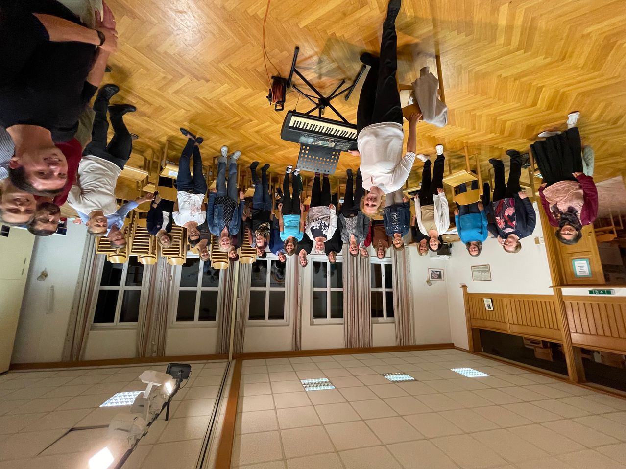 Eine Gruppe von Menschen steht kopfüber in einem Raum mit Holzboden, wahrscheinlich für ein Foto. Es gibt ein Klavier auf einem Ständer und einen Spiegel auf der linken Seite.