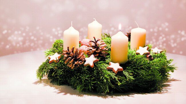 Ein Kranz mit vier brennenden Kerzen, Tannenzapfen und Sternen auf einem weißen Tisch vor einem rosa Hintergrund.