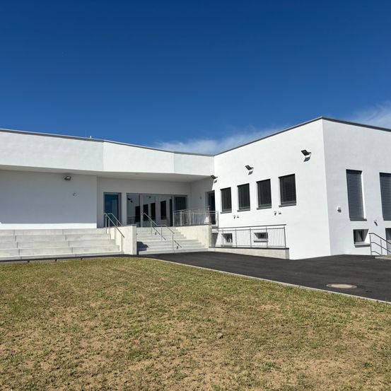 Ein modernes weißes Gebäude mit flachem Dach, mehreren Fenstern und blauem Himmel im Hintergrund. Es gibt Treppen, die zum Eingang führen, mit Geländern an den Seiten.