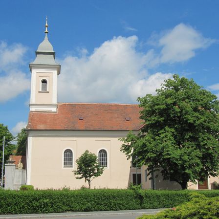 Eine weiße Kirche mit einem roten Dach und einem hohen Turm steht unter einem blauen Himmel mit Wolken. Davor stehen Büsche.