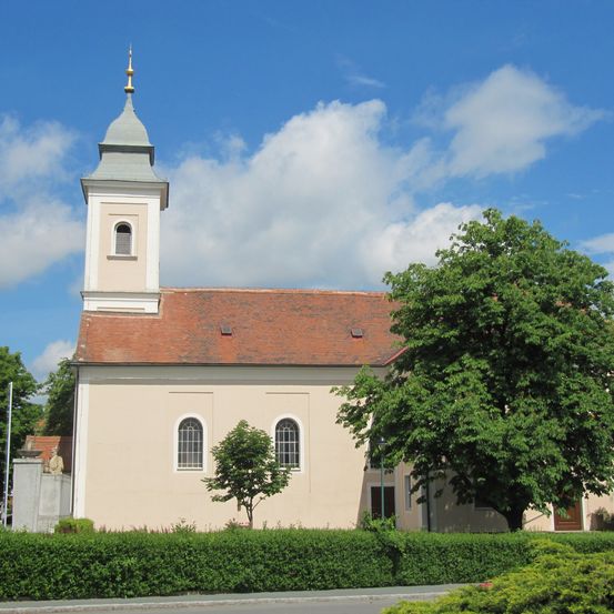 Eine weiße Kirche mit einem roten Dach und einem hohen Turm steht unter einem blauen Himmel mit Wolken. Davor stehen Büsche.