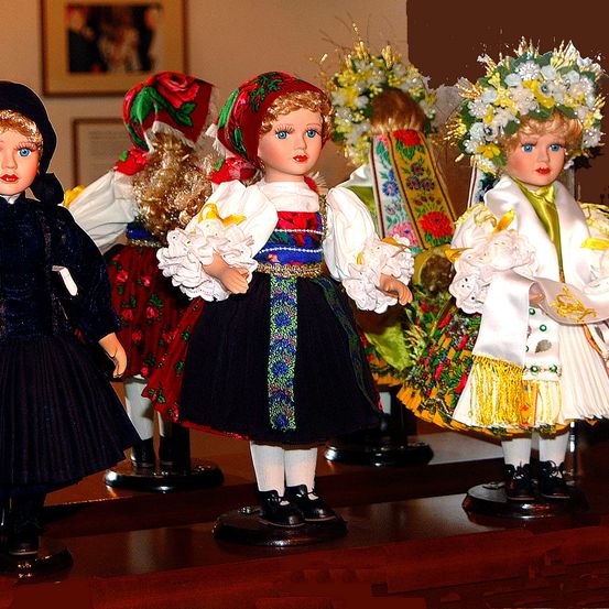 Fünf Puppen in traditioneller Kleidung sind auf einem Holztisch ausgestellt. Jede Puppe hat eine unterschiedliche Kleidung, einschließlich Blumenkränze und detailreiche Kleider. Im Hintergrund befinden sich gerahmte Bilder an der Wand.