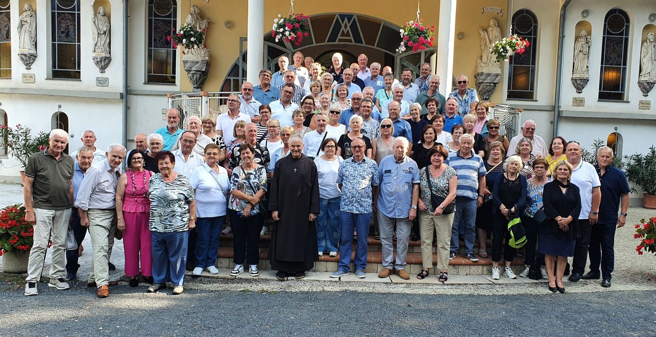 Eine Gruppe von Menschen, darunter ältere Erwachsene, steht auf den Stufen eines Gebäudes mit dekorativen Bögen und Blumen. Ein Priester steht davor, und sie alle lächeln für ein Foto.