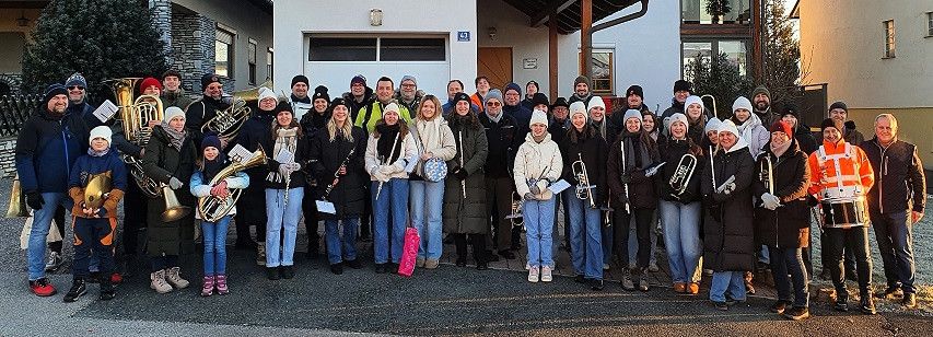 Eine Gruppe von Menschen, einschließlich Kindern, posiert für ein Foto vor einem Gebäude. Sie halten verschiedene Musikinstrumente wie Trompeten, Posaunen und Trommeln.