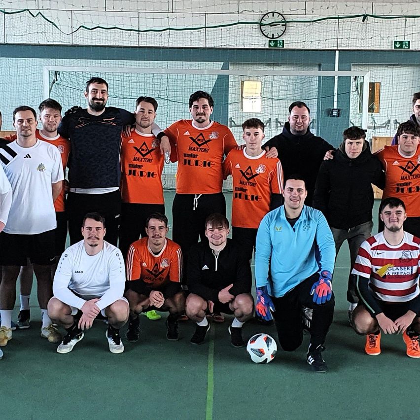 Eine Gruppe von Fußballspielern posiert für ein Foto vor einem Tor mit orangefarbenen Trikots, ein Spieler trägt eine blaue Jacke und ein Fußball liegt auf dem Boden.