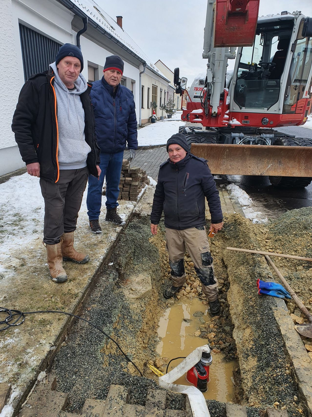 Drei Männer stehen an einem verschneiten Tag draußen in der Nähe einer Grube. Ein Mann ist in der Grube und hält ein Werkzeug. Hinter ihnen steht eine rot-weiße Maschine.