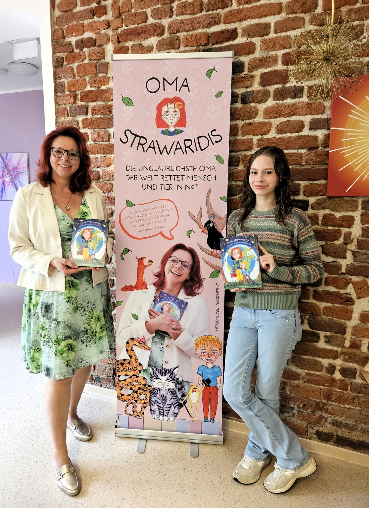 Zwei Frauen stehen neben einem Buchbanner. Die Frau auf der linken Seite hält ein Buch und lächelt. Die Frau auf der rechten Seite hält ein Buch mit einer Illustration eines Kindes und einer Katze. Ein Backsteinmauer steht hinter ihnen.