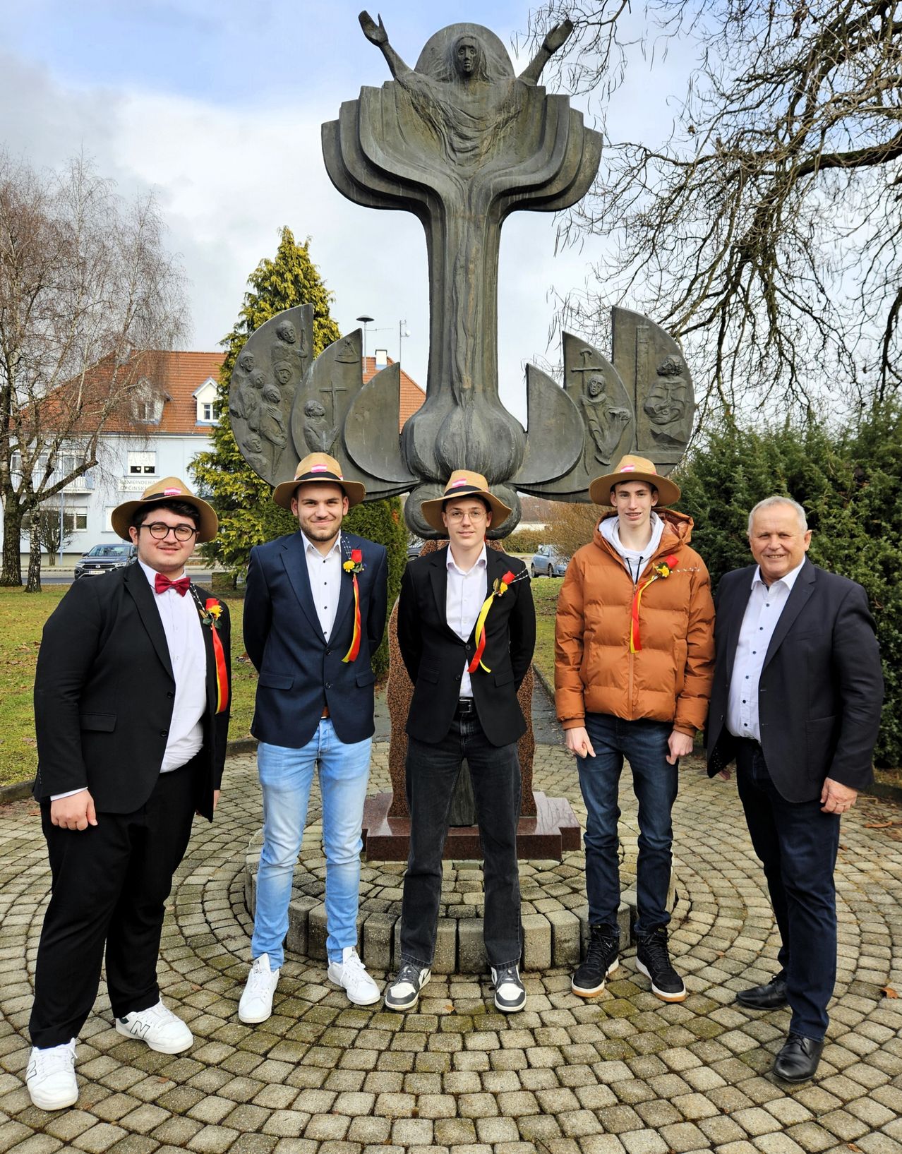 Five men stand before a large, abstract statue. They wear formal attire, including hats and bow ties. Behind them, a house and trees are visible.