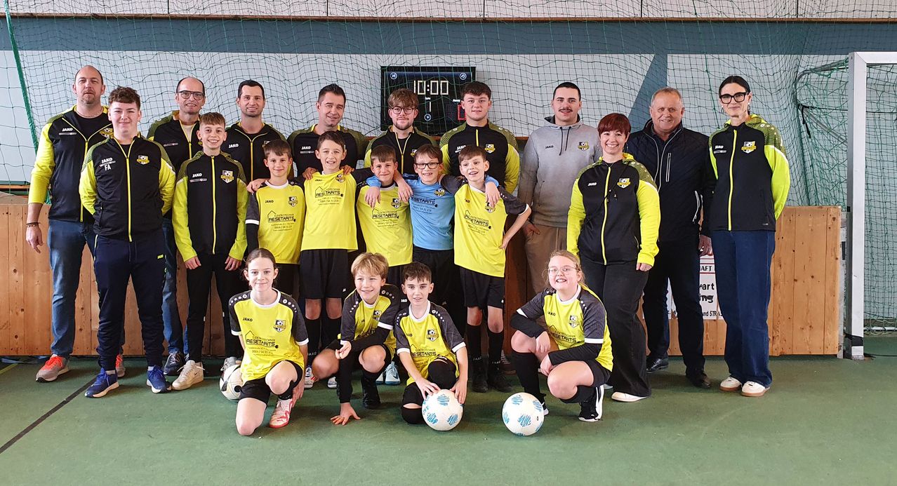 A soccer team wearing yellow and black uniforms poses for a group photo in front of a net. The team includes children and adults, with a digital clock in the background showing the time as 10:00.