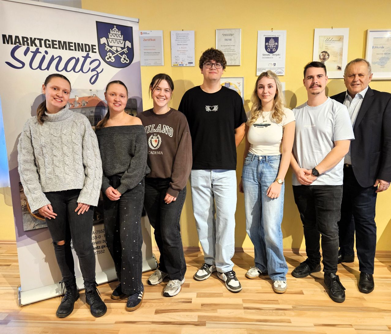 Eine Gruppe junger Erwachsener steht lächelnd zusammen auf einem Holzboden. Sie tragen Freizeitkleidung und scheinen für ein Foto zu posieren. Hinter ihnen hängt eine gelbe Wand mit mehreren gerahmten Dokumenten. Ein Banner mit Logo und Text befindet sich auf der linken Seite.