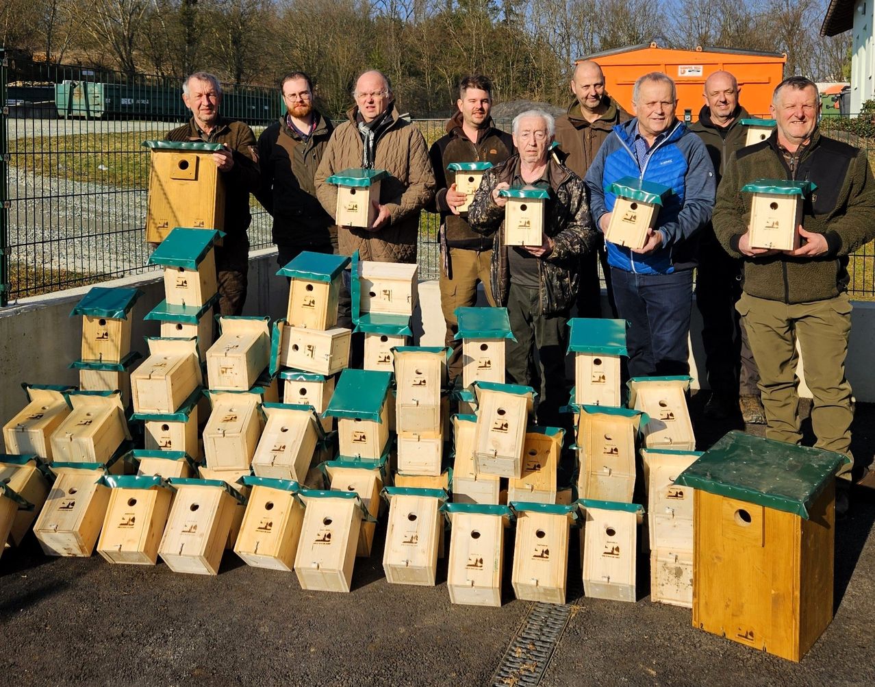 Eine Gruppe von Männern posiert mit mehreren Vogelhäuschen vor einem Zaun mit Bäumen und einer orangefarbenen Struktur im Hintergrund.