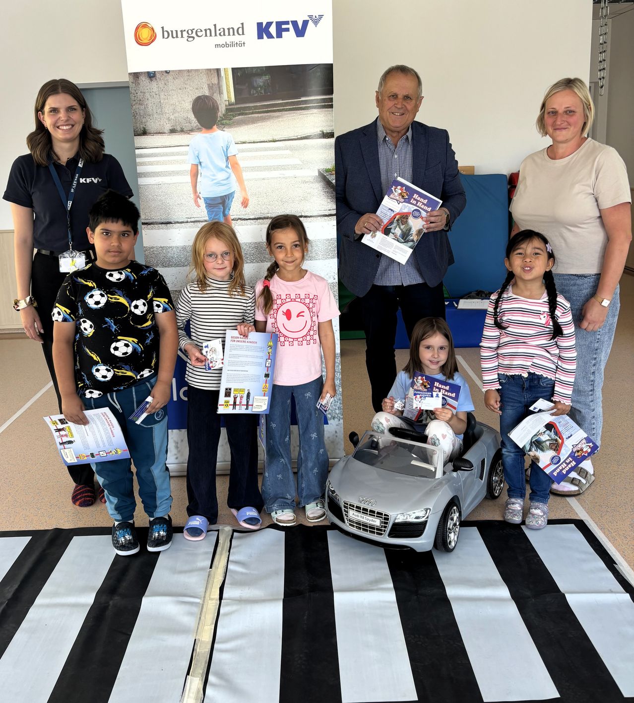 Eine Gruppe von Erwachsenen und Kindern steht zusammen in einem Raum, einige halten Bücher und posieren für ein Foto. Ein Kind in einem Spielzeugauto, ein Zebrastreifen-Banner im Hintergrund.