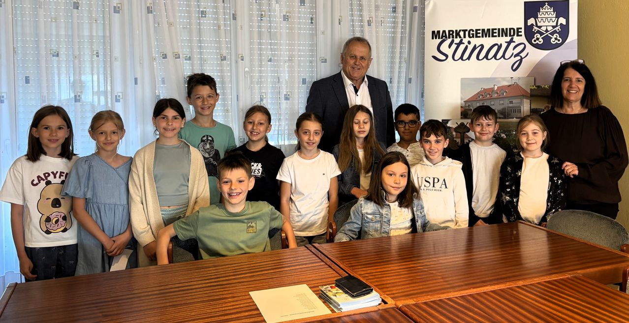 Eine Gruppe von Kindern vor einem Schreibtisch mit einem Mann im Anzug. Sie lächeln und scheinen für ein Foto zu posieren. Dahinter ist ein Banner mit den Worten 'MARKTGE Sti'.
