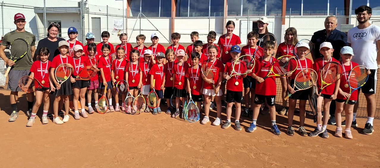 A group of young tennis players, all wearing red shirts and holding tennis rackets, stand on a tennis court with a blurry background.