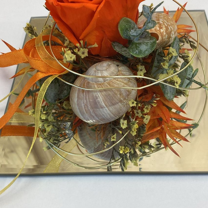 Eine dekorative Anordnung mit einer orangefarbenen Rose, Sukkulenten und Muscheln auf einer goldplattierten Basis. Das Blumenarrangement besteht aus gelben und orangefarbenen Blättern und kleinen Blüten.