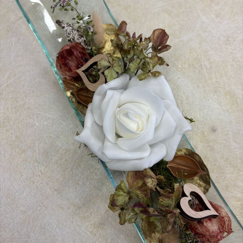 Eine klare Glasvase gefüllt mit einer weißen Rose, getrockneten Blättern und Holzherzen, die auf einer strukturierten Oberfläche ruht.