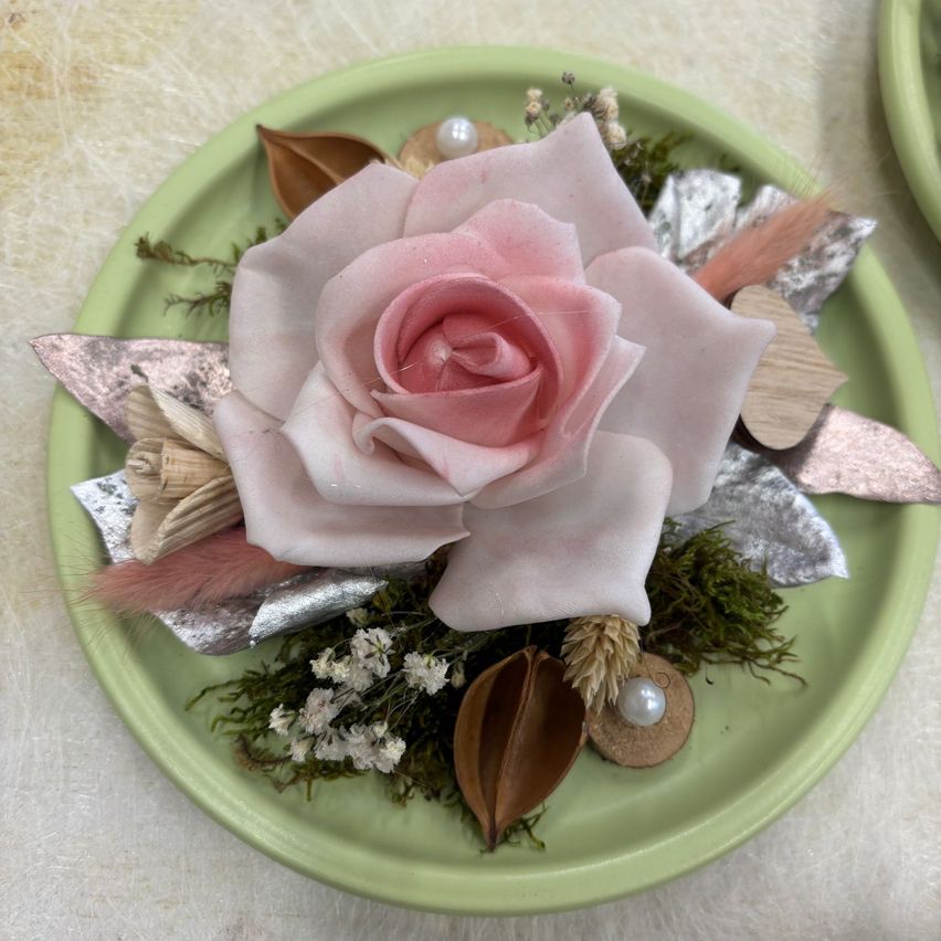 Ein grüner Teller mit einer rosa Rose, Perlen, Federn und Moos auf einer weißen Oberfläche.