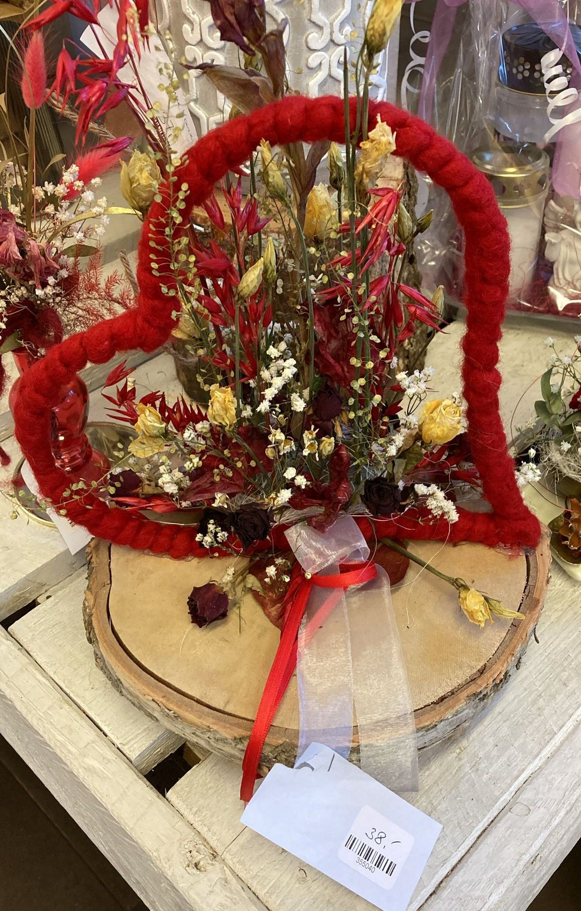 Ein herzförmiges Blumenarrangement mit roten und gelben Blumen auf einem Holzsockel, gebunden mit einem roten Band. Andere Blumen und Kerzen sind im Hintergrund.