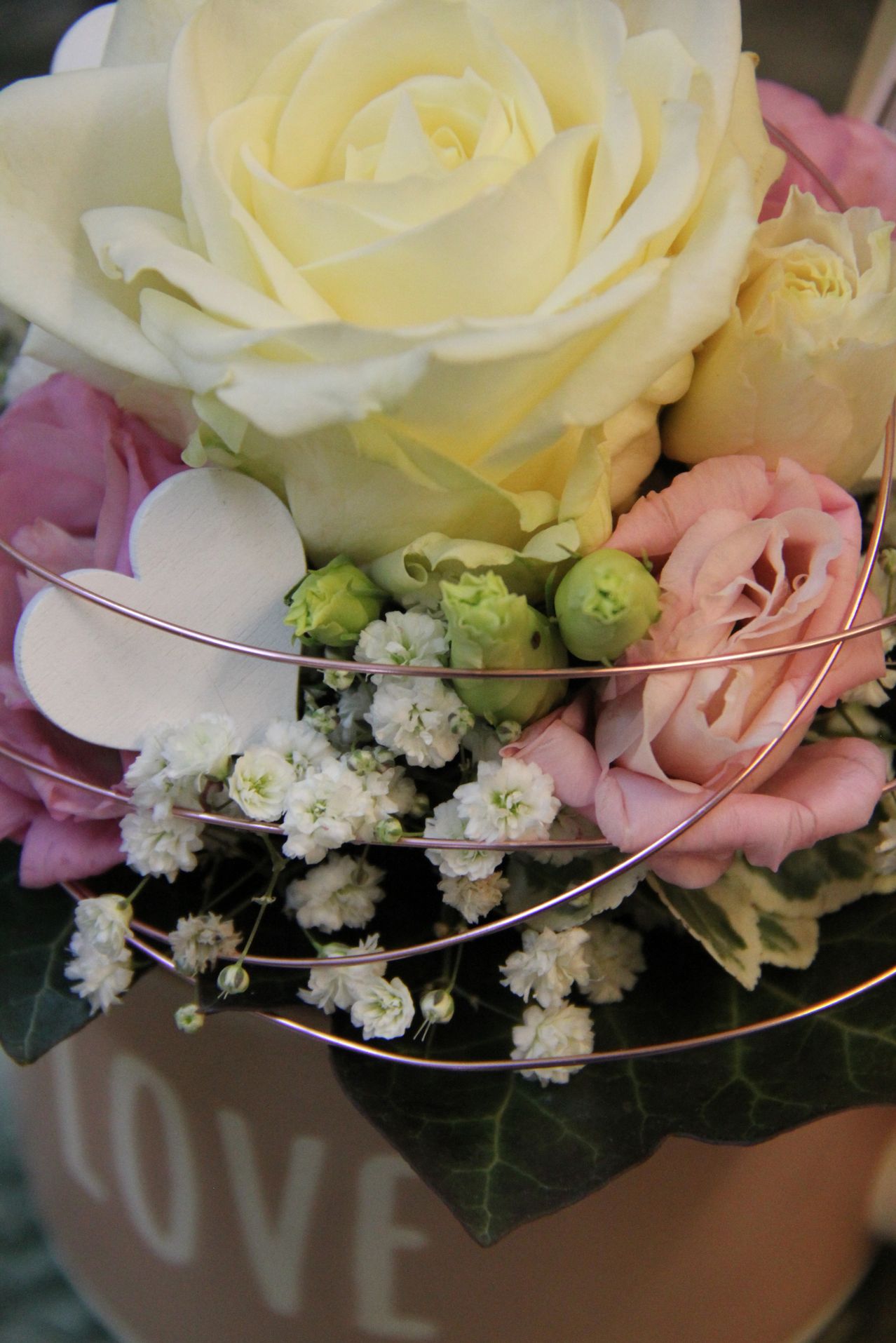 A close-up view of a floral arrangement features a large white rose, surrounded by pink roses and white baby's breath. A heart-shaped wooden piece and metallic wires hold the flowers together.