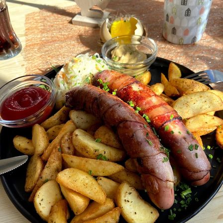 Ein schwarzer Teller mit zwei in Speck gewickelten Würsten, knusprigen Kartoffeln und verschiedenen Beilagen wie Ketchup und Dip. Eine Gabel und ein Messer liegen in der Nähe.