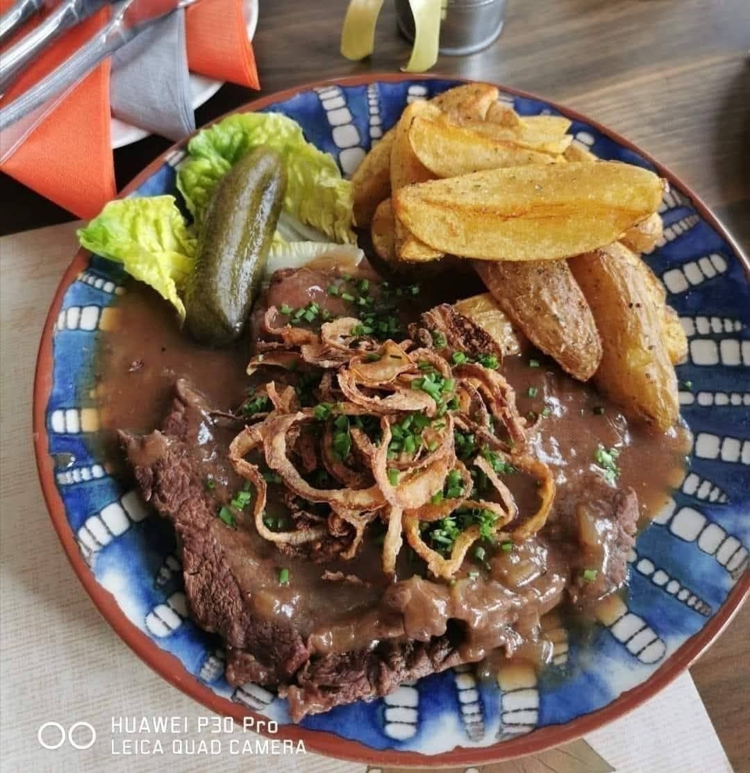 Ein Teller mit Steak in Soße, gebratenen Zwiebeln und Petersilie, begleitet von einem Gewürzgurke, Salat und Pommes frites.