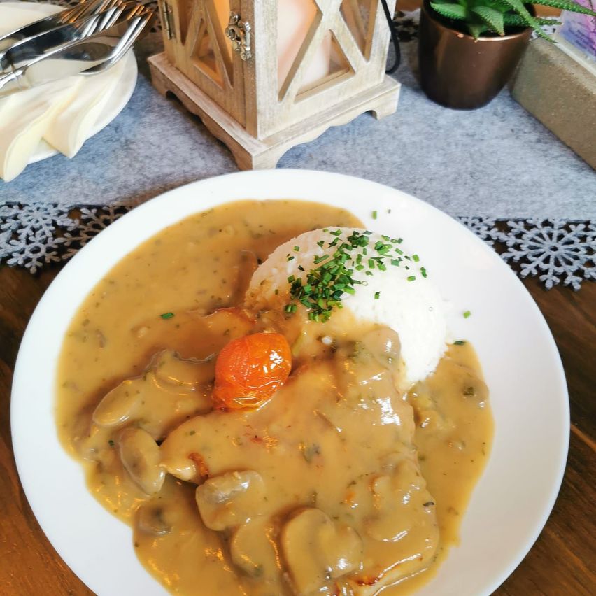 Ein Teller mit cremigem Champignonhähnchen mit Reisbeilage, garniert mit einer Kirschtomate und Petersilie. Auf einem Holztisch mit einer Kerze und einer Pflanze im Hintergrund serviert.