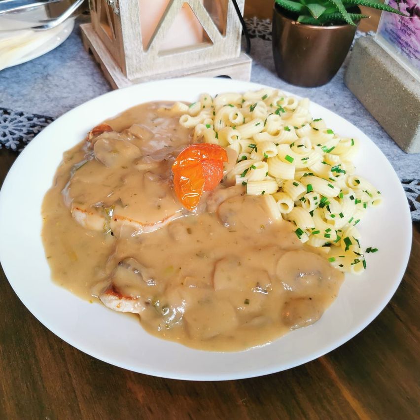 Ein weißer Teller mit einem Schweinekotelett, bedeckt mit Pilzsoße und Nudeln, mit einer Scheibe Tomate auf der Seite.