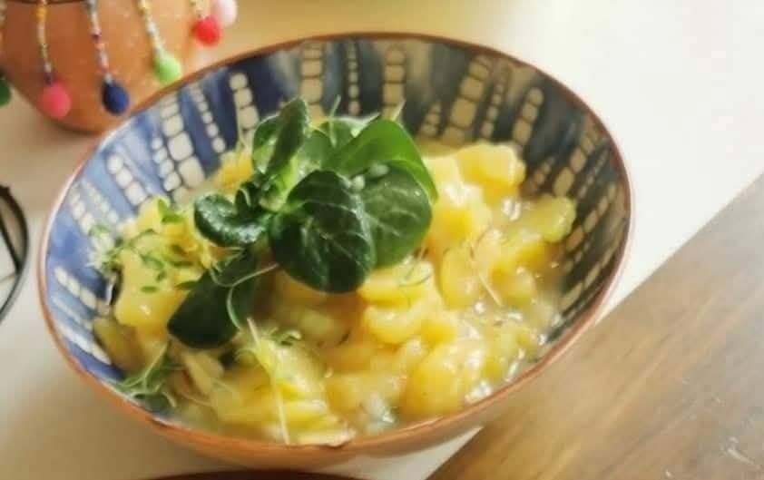 A bowl of creamy potato salad with green herbs on top, placed on a wooden table.
