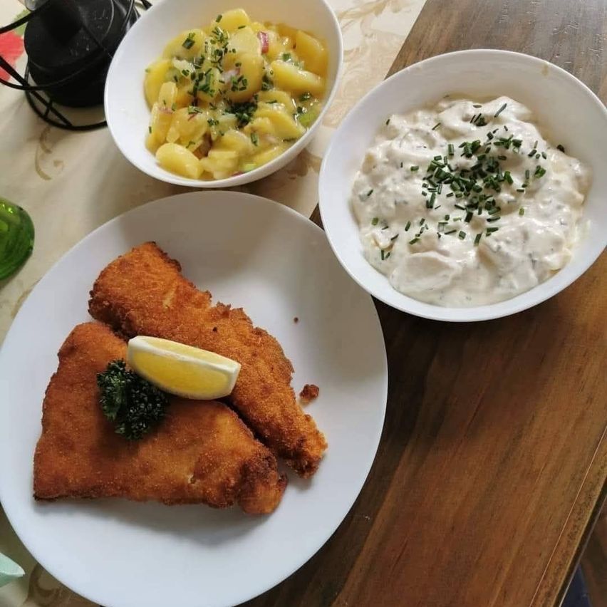 Auf einem Holztisch steht ein Teller mit gebratenem Fisch, einer Zitronenscheibe und Brokkoli, begleitet von einer Schüssel Kartoffelsalat und einer Schüssel cremiger Sauce.