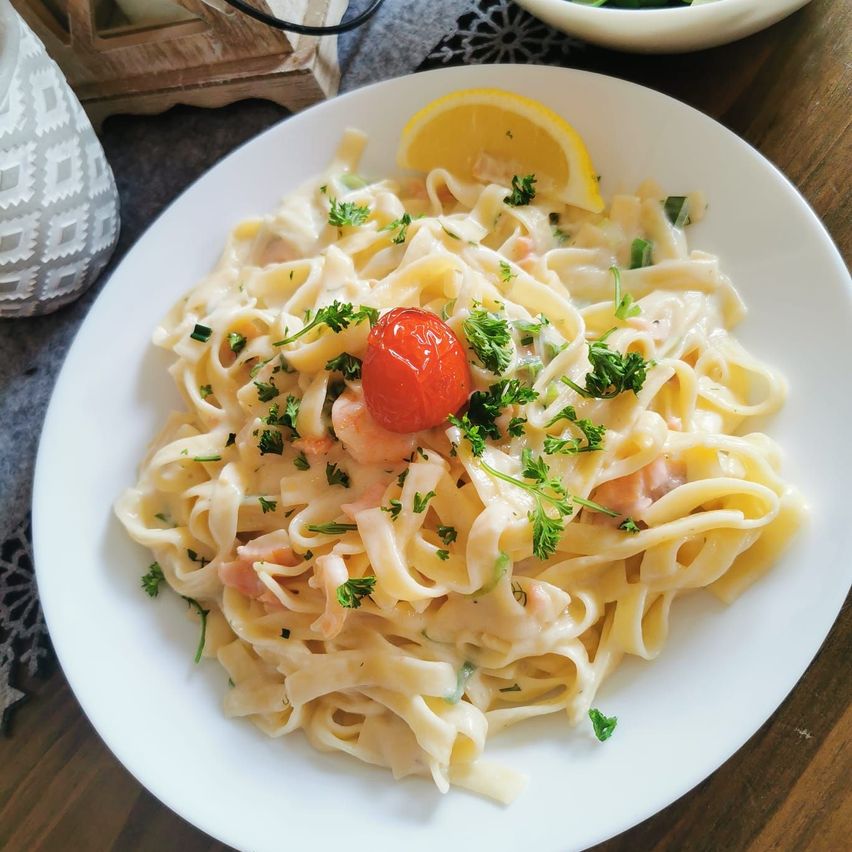 Ein weißer Teller mit cremiger Pasta, garniert mit Garnelen, einer Kirschtomate und Petersilie. Eine Zitronenscheibe befindet sich neben dem Teller.
