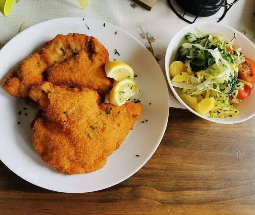 Ein weißer Teller mit paniertem Fleisch, zwei Zitronenscheiben und einer kleinen Schüssel Salat auf einem Holztisch.