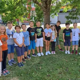 Eine Gruppe von Kindern und Jugendlichen posiert für ein Foto unter einem Baum in einem Rasenbereich. Hinter ihnen befinden sich Gebäude und ein geparktes Auto.