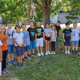 A group of children and young adults posing for a picture under a tree in a grassy area. Behind them are buildings and a parked car.