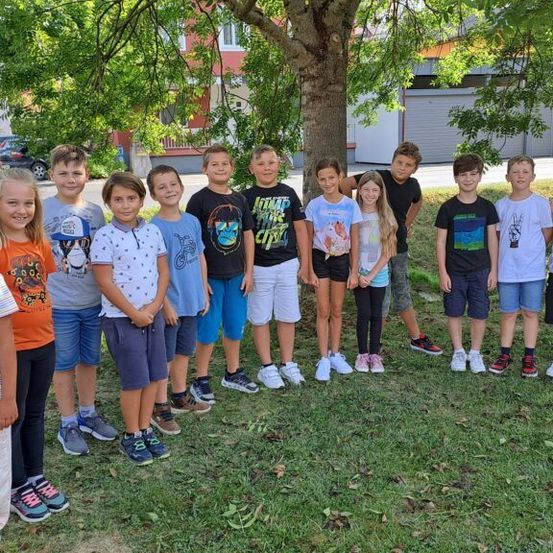 Eine Gruppe von Kindern und Jugendlichen posiert für ein Foto unter einem Baum in einem Rasenbereich. Hinter ihnen befinden sich Gebäude und ein geparktes Auto.