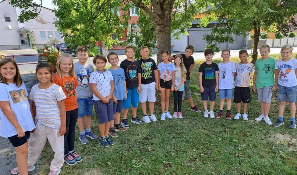 Eine Gruppe von Kindern und Jugendlichen posiert für ein Foto unter einem Baum in einem Rasenbereich. Hinter ihnen befinden sich Gebäude und ein geparktes Auto.