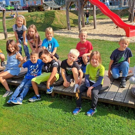 Eine Gruppe von Kindern sitzt auf einer Holzplattform in einem Spielplatz. Einige tragen Turnschuhe, während andere Sandalen haben. Hinter ihnen befindet sich eine rote Rutsche.
