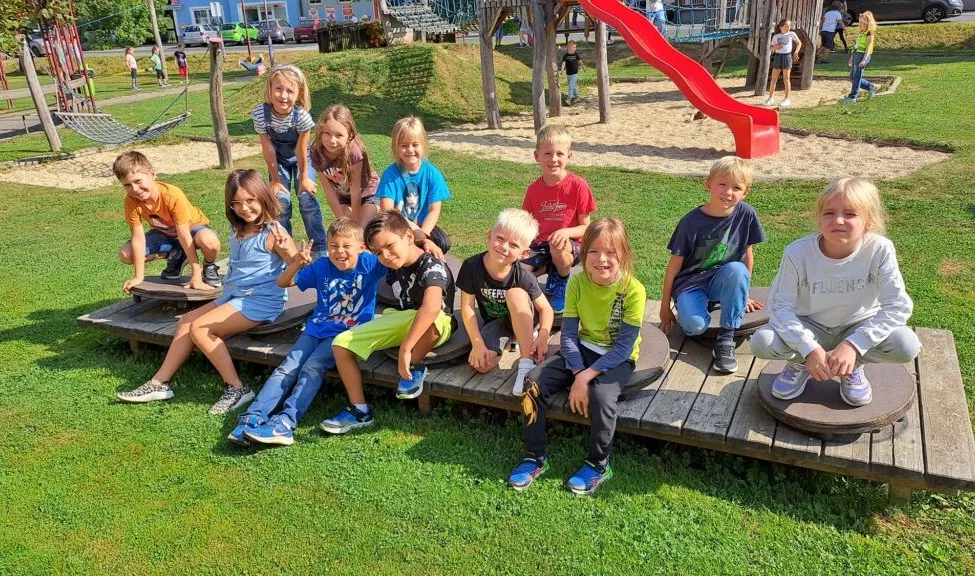 Eine Gruppe von Kindern sitzt auf einer Holzplattform in einem Spielplatz. Einige tragen Turnschuhe, während andere Sandalen haben. Hinter ihnen befindet sich eine rote Rutsche.