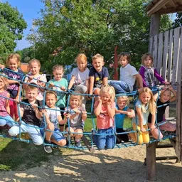 Eine Gruppe von Kindern spielt auf einer blauen Strickleiter in einem Park unter einem sonnigen Himmel. Sie lächeln und posieren für ein Foto.