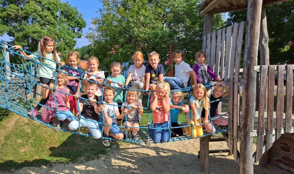 Eine Gruppe von Kindern spielt auf einer blauen Strickleiter in einem Park unter einem sonnigen Himmel. Sie lächeln und posieren für ein Foto.