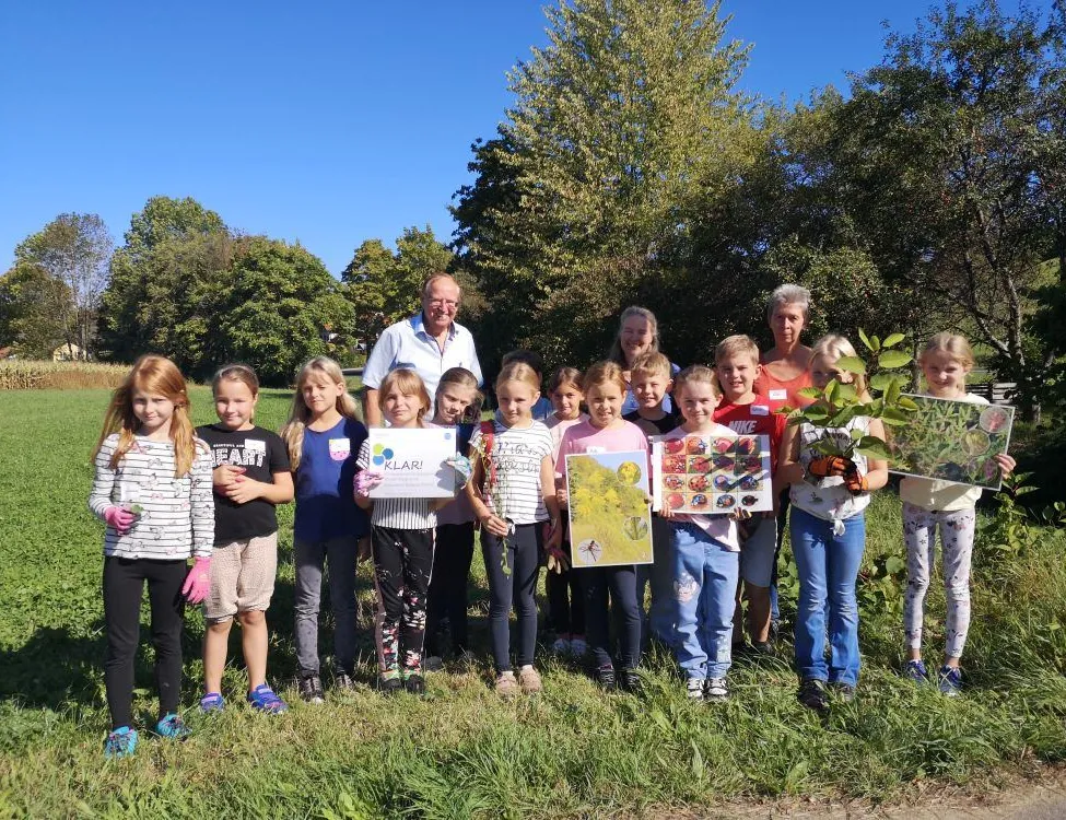 Eine Gruppe junger Kinder und Erwachsener posiert für ein Foto in einem Park mit Bäumen im Hintergrund. Die Kinder halten Pflanzen und Schilder in den Händen.