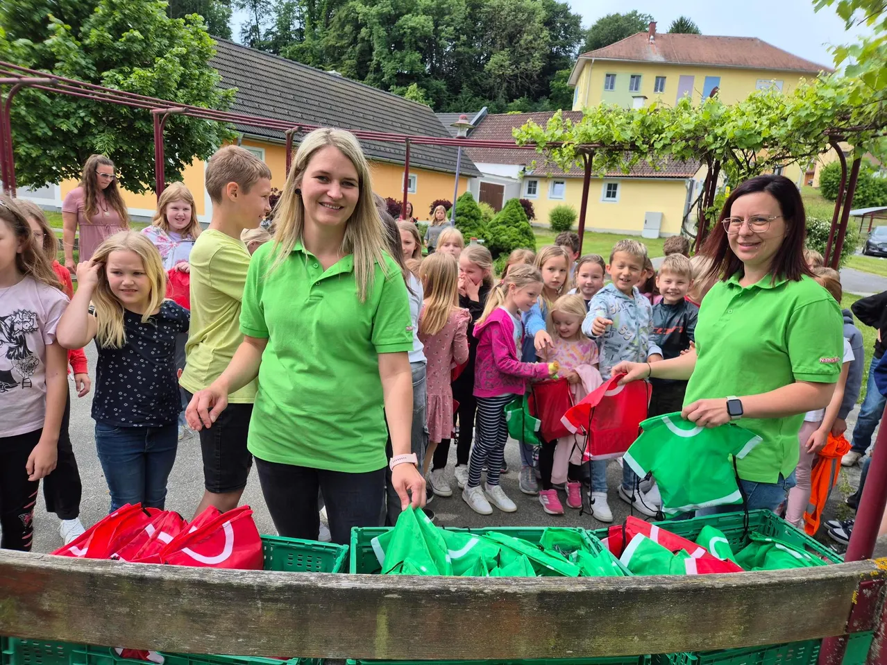 Eine Gruppe von Kindern und Erwachsenen in grünen Shirts halten rote und grüne Taschen in einem Außenbereich.