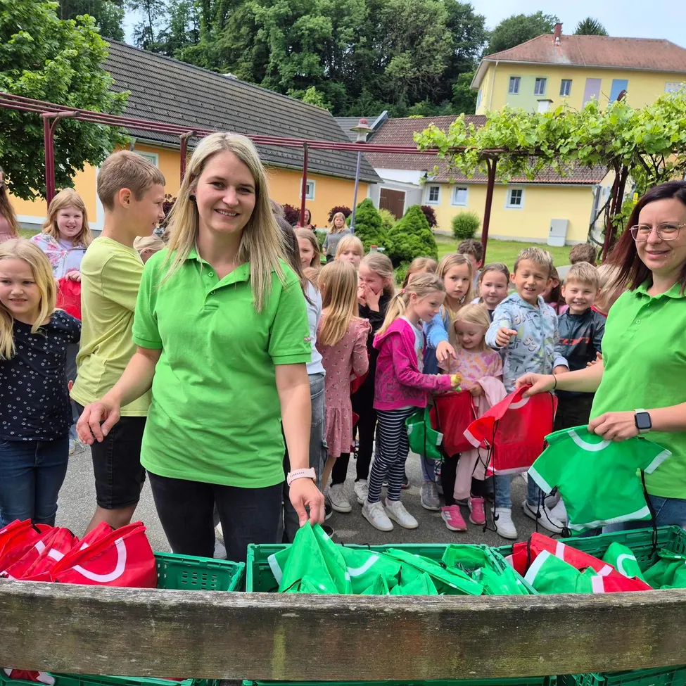 Eine Gruppe von Kindern und Erwachsenen in grünen Shirts halten rote und grüne Taschen in einem Außenbereich.