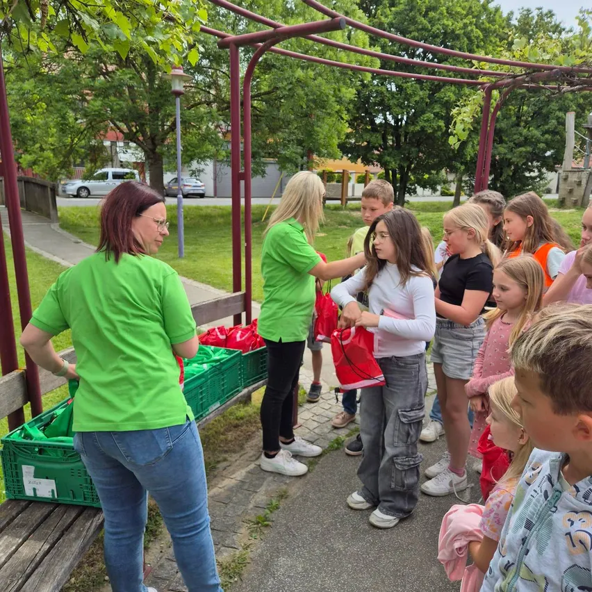 Mehrere Erwachsene und Kinder in grünen T-Shirts versammeln sich um eine Frau, die einen roten Beutel hält. Sie stehen in einem Park mit grünen Kisten, Bäumen und einem Weg.