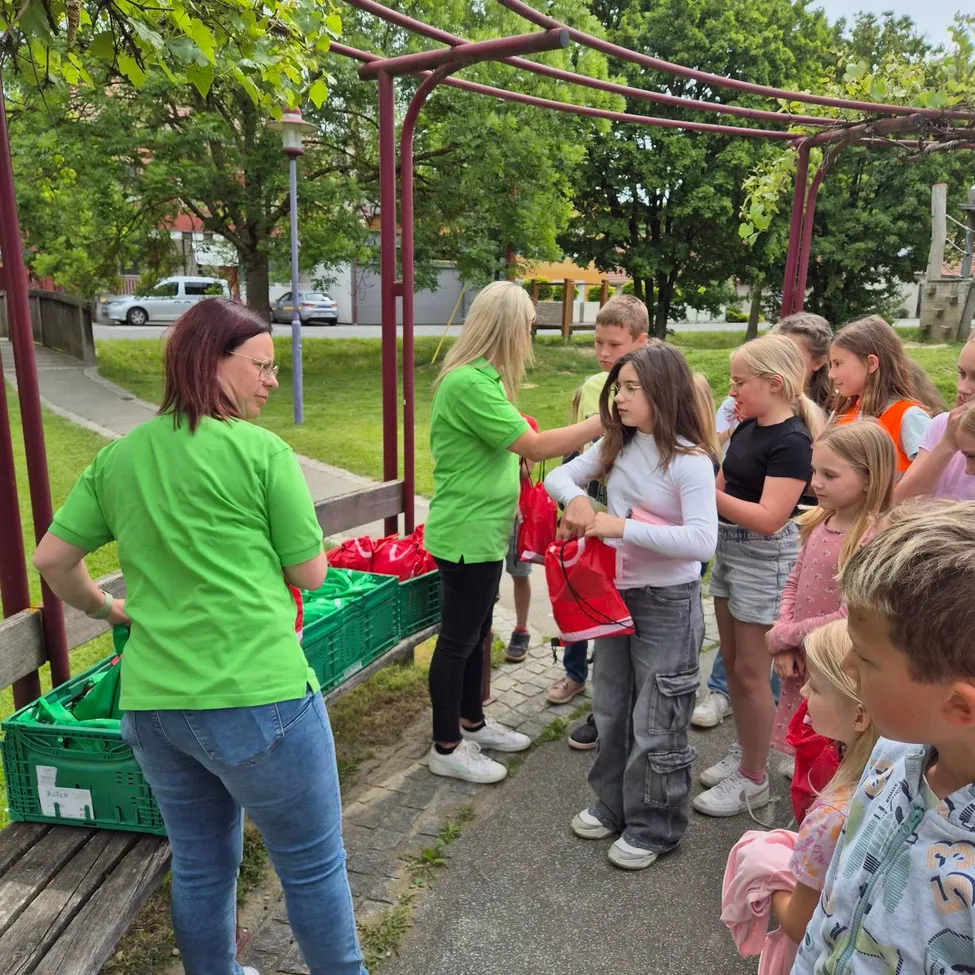 Mehrere Erwachsene und Kinder in grünen T-Shirts versammeln sich um eine Frau, die einen roten Beutel hält. Sie stehen in einem Park mit grünen Kisten, Bäumen und einem Weg.