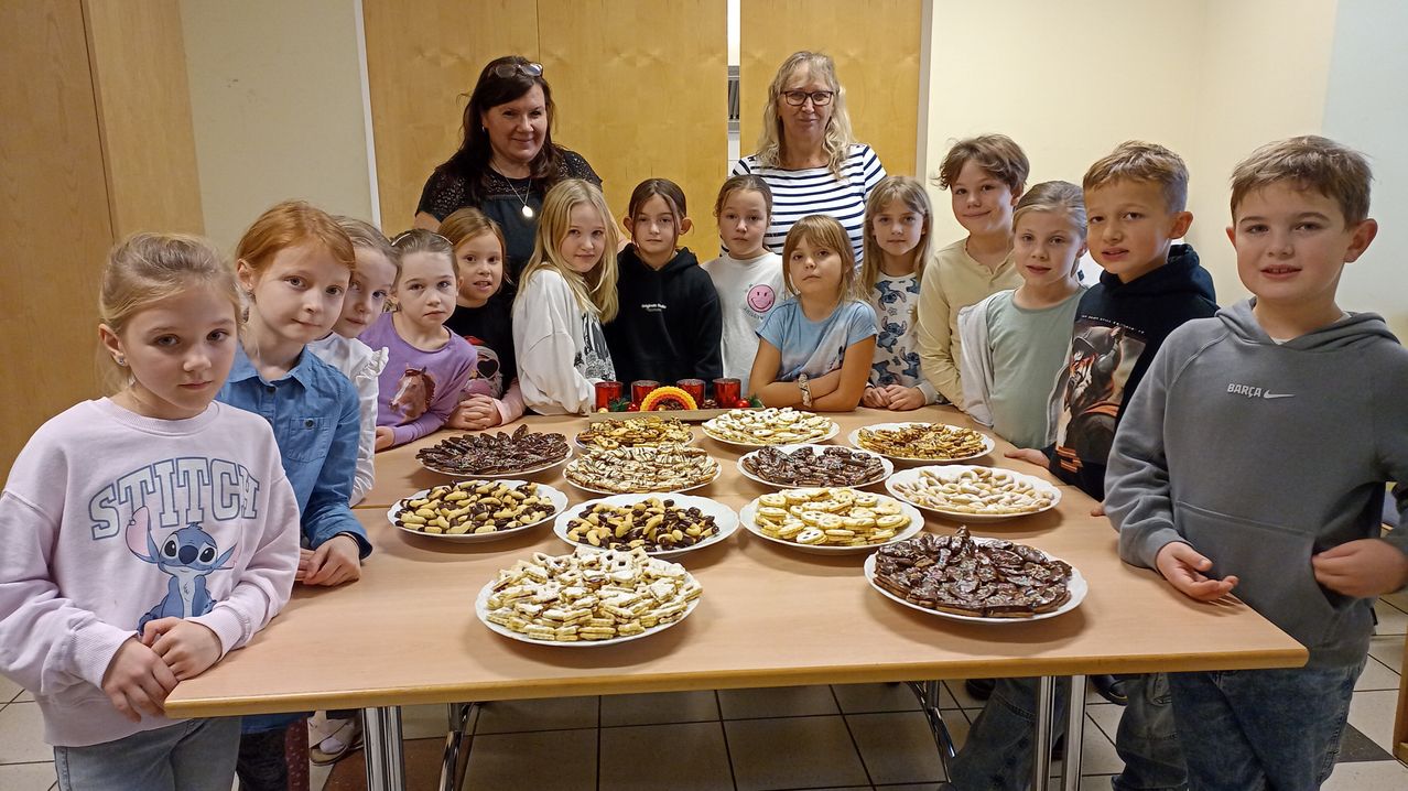 Eine Gruppe von Kindern und zwei Frauen stehen hinter einem Tisch voller verschiedener Arten von Keksen. Die Kekse stehen auf weißen Tellern und sind in Reihen angeordnet. Die Frauen tragen Brillen und Halsketten.