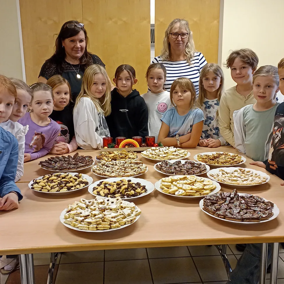Eine Gruppe von Kindern und zwei Frauen stehen hinter einem Tisch voller verschiedener Arten von Keksen. Die Kekse stehen auf weißen Tellern und sind in Reihen angeordnet. Die Frauen tragen Brillen und Halsketten.